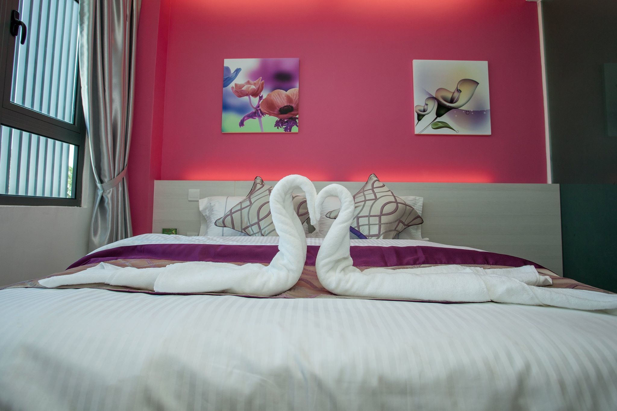 Un lit bien fait, avec du linge de lit blanc et deux oreillers, un mur rose orné de deux tableaux floraux et une fenêtre avec des rideaux.
