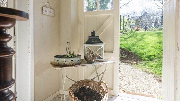 Un interior rústico con una puerta blanca que indica “JARDIN”, una pequeña mesa con un farol y una maceta rústica, y una cesta de mimbre llena de piñas.
