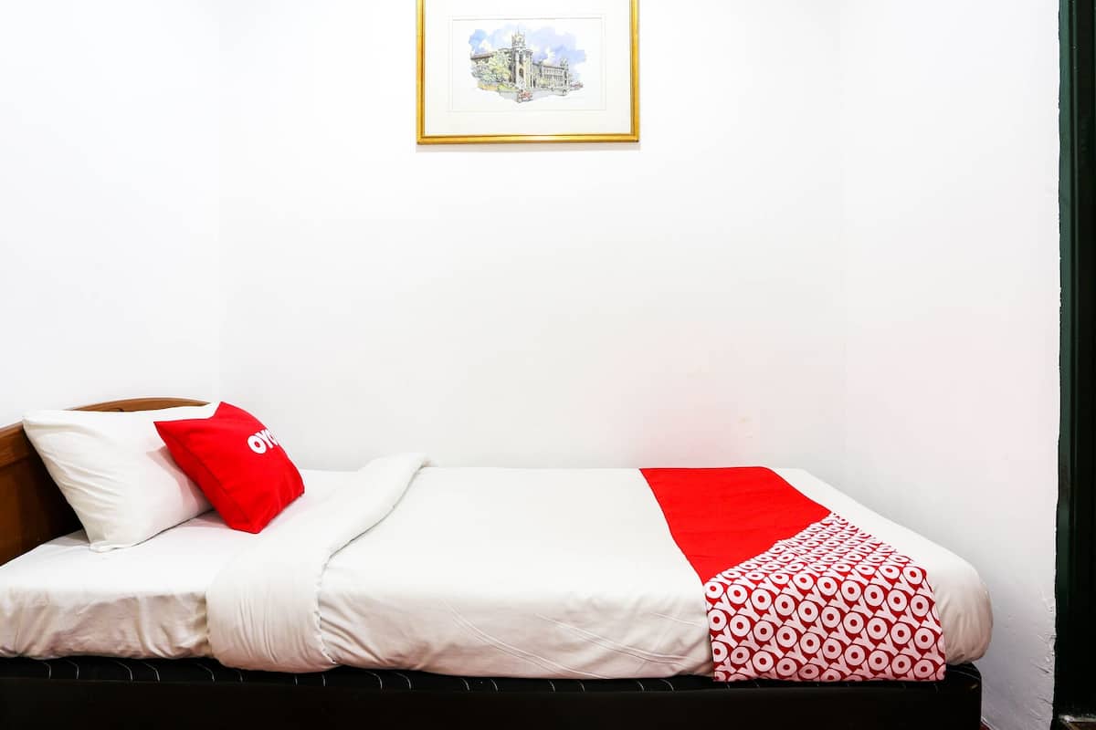 Una cama individual con ropa de cama blanca y una manta estampada en rojo y blanco. Encima de la cama, hay un cuadro enmarcado colgado en la pared.
