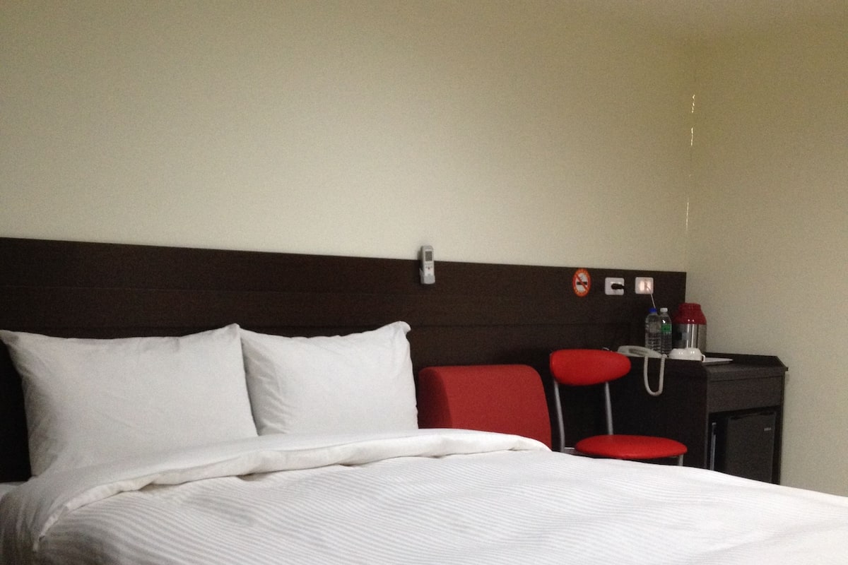 A hotel room with a large bed, a bedside table with a phone and a water bottle, and a red chair.