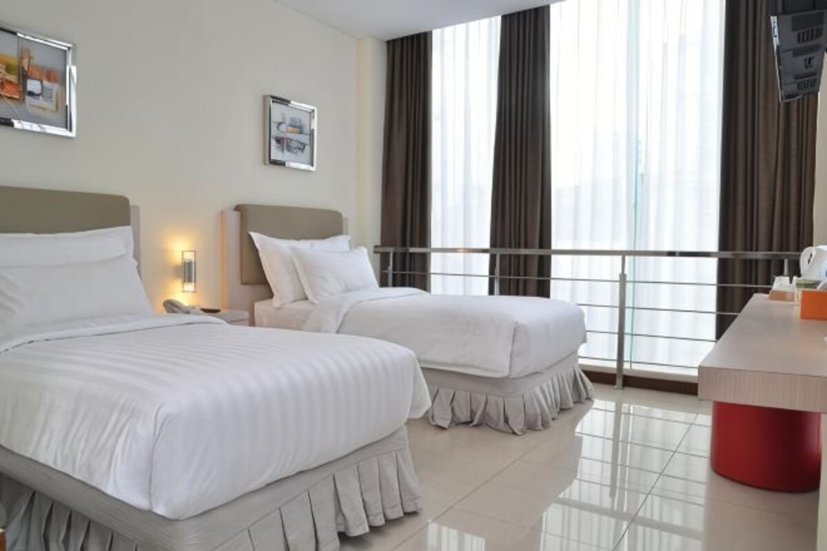 Habitación de hotel con dos camas, un escritorio, un televisor y ventanas grandes con cortinas.