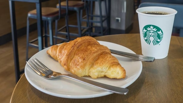 Un croissant en un plato con un tenedor y un cuchillo, y una taza Starbucks sobre una mesa en un café.