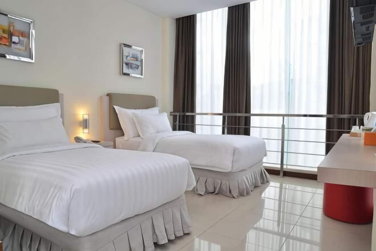 Habitación de hotel con dos camas, un escritorio, un televisor y ventanas grandes con cortinas.