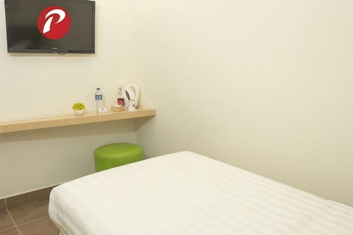 A hotel room with a bed, a small table with a kettle, a TV, and a green trash bin.