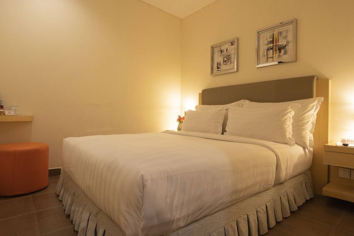 Ein ordentlich bezogenes Bett mit Kopfteil, ein Nachttisch mit Lampe und ein kleiner orangefarbener Hocker.