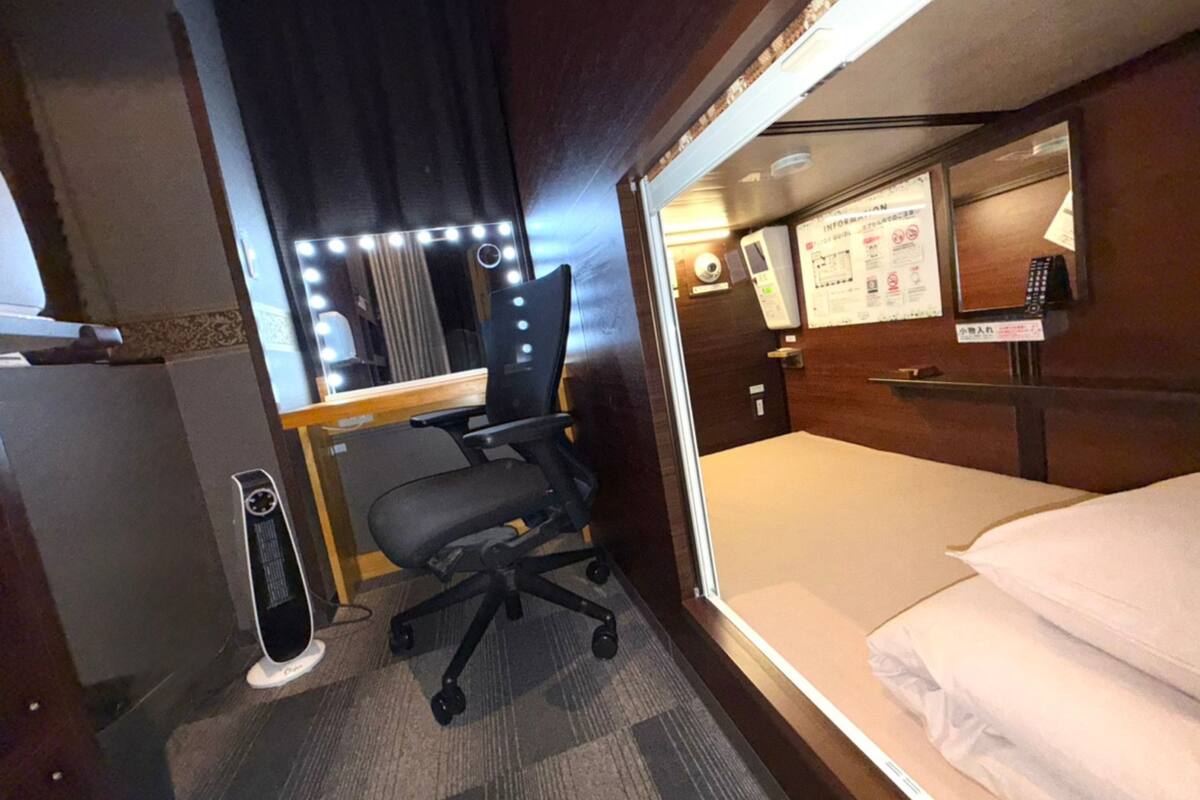 Een kleine hotelkamer met een bed, een bureau met stoel, een spiegel met verlichting, een klein verwarmingstoestel en een tv aan de muur.