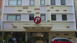 Un bâtiment moderne, surmonté d’une enseigne indiquant « d’primahotel ». Deux voitures sont garées devant, et l’entrée est dotée d’une porte en verre.