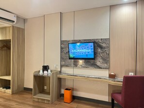 Zona de recepción de hotel con un televisor montado en una pared de mármol, un armario de madera, un rincón de descanso con una silla roja y un pequeño cubo de basura.