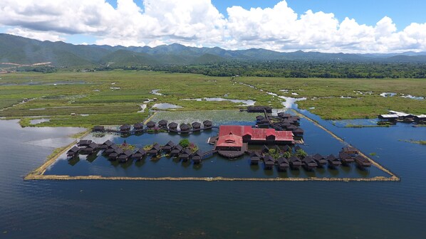 Eine Vogelperspektive auf ein schwimmendes Hotel mit überwasserbungalows, die durch Wege miteinander verbunden sind, umgeben von üppigen grünen Feldern und Bergen im Hintergrund.