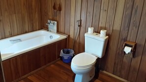 Ein kleines, kompaktes Badezimmer mit Holzvertäfelung, einer weißen Badewanne, einer Toilette und einem blauen Mülleimer.