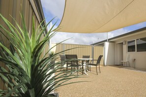 Um terraço ao ar livre com mesa e cadeiras, uma planta grande e um edifício ao fundo.