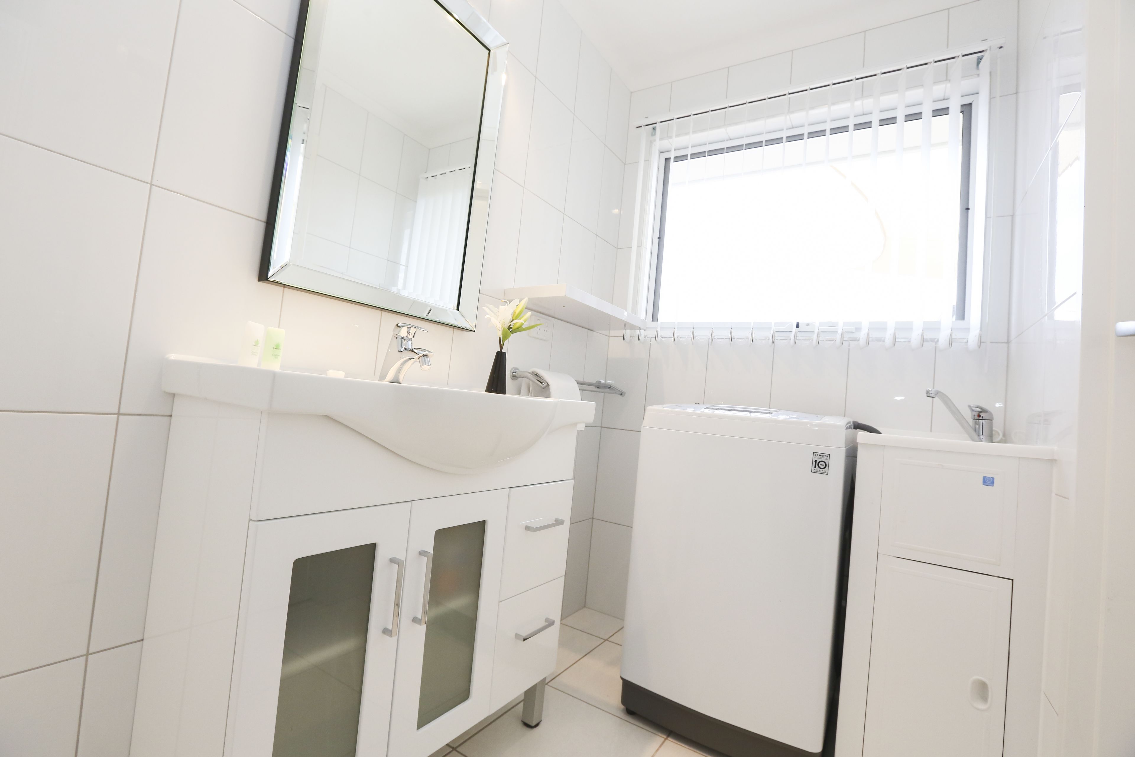 A modern bathroom with a white sink, mirror, and a window with blinds.