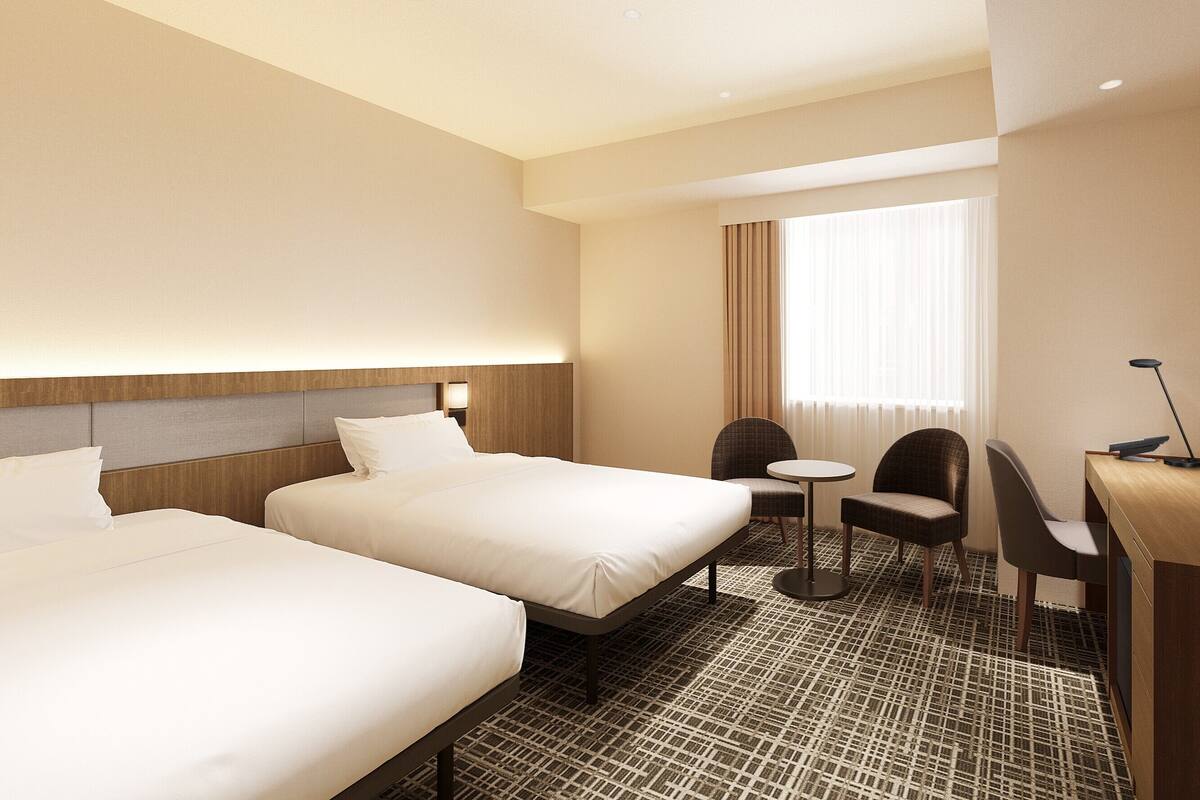 Une chambre d’hôtel avec deux lits, une petite table ronde, deux chaises, un bureau avec une lampe et une grande fenêtre avec des rideaux.