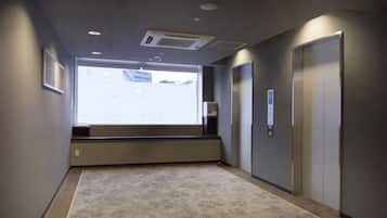 A hallway with a carpeted floor, a window with blinds, an air vent, and a wall-mounted elevator control panel.
