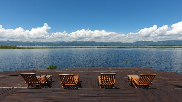 A wooden deck with four wooden chairs facing a calm lake, surrounded by mountains and a clear sky. - Golden Island Cottages Thale-U Hotel (Nyaungshwe)