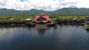 A traditional houseboat with a red roof, wooden deck, and a small porch. It is situated on a calm body of water with lush green hills in the background. - Golden Island Cottages Thale-U Hotel (Nyaungshwe)