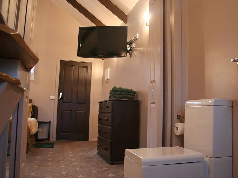Un pasillo estrecho con una puerta de madera oscura, un inodoro blanco, un refrigerador pequeño y blanco, un aparador de madera oscura y un televisor de pantalla plana montado en la pared.
