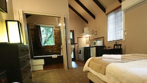 A bedroom with a bed, a dresser, a desk with a chair, a window with blinds, and a stone wall.