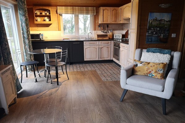 A compact kitchen with wooden cabinets, a dining area with a table and chairs, and a cozy seating area with a cushioned chair. - Cottage in Brookland with Private Sauna (Fairfield)