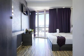 Una habitación de hotel moderna con una cama, televisión, escritorio con planta y un amplio ventanal.