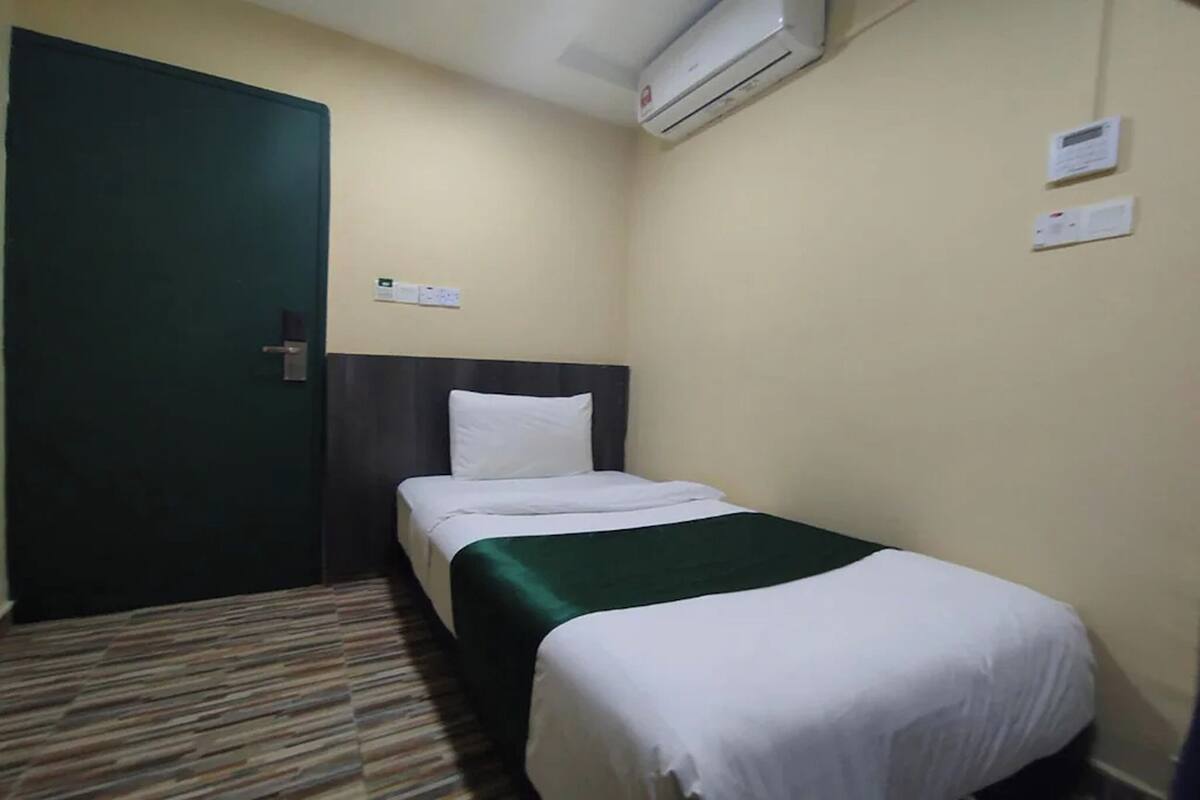 A single bed with a green bedspread, a dark headboard, a white pillow, and an air conditioning unit mounted on the wall.