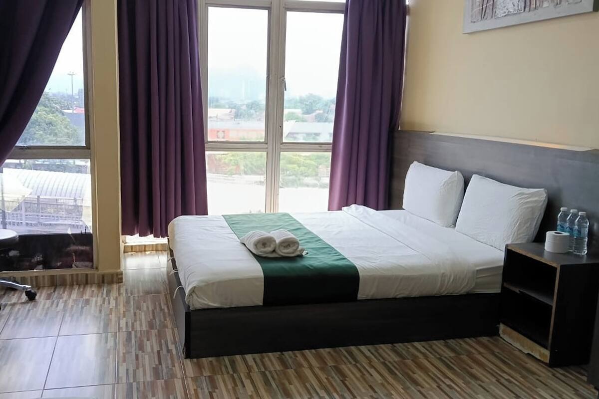 A hotel room with a bed, a nightstand, a window with curtains, and a view of the cityscape.