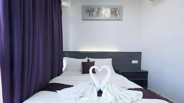 Une chambre d’hôtel avec un lit, une table de chevet, une œuvre d’art accrochée au mur et des rideaux violets.