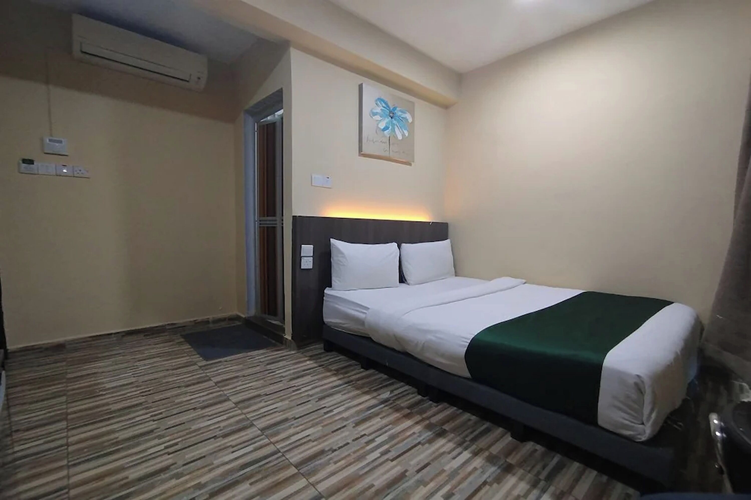 A hotel room with a bed, an air conditioning unit, a door, and a wall-mounted light fixture.