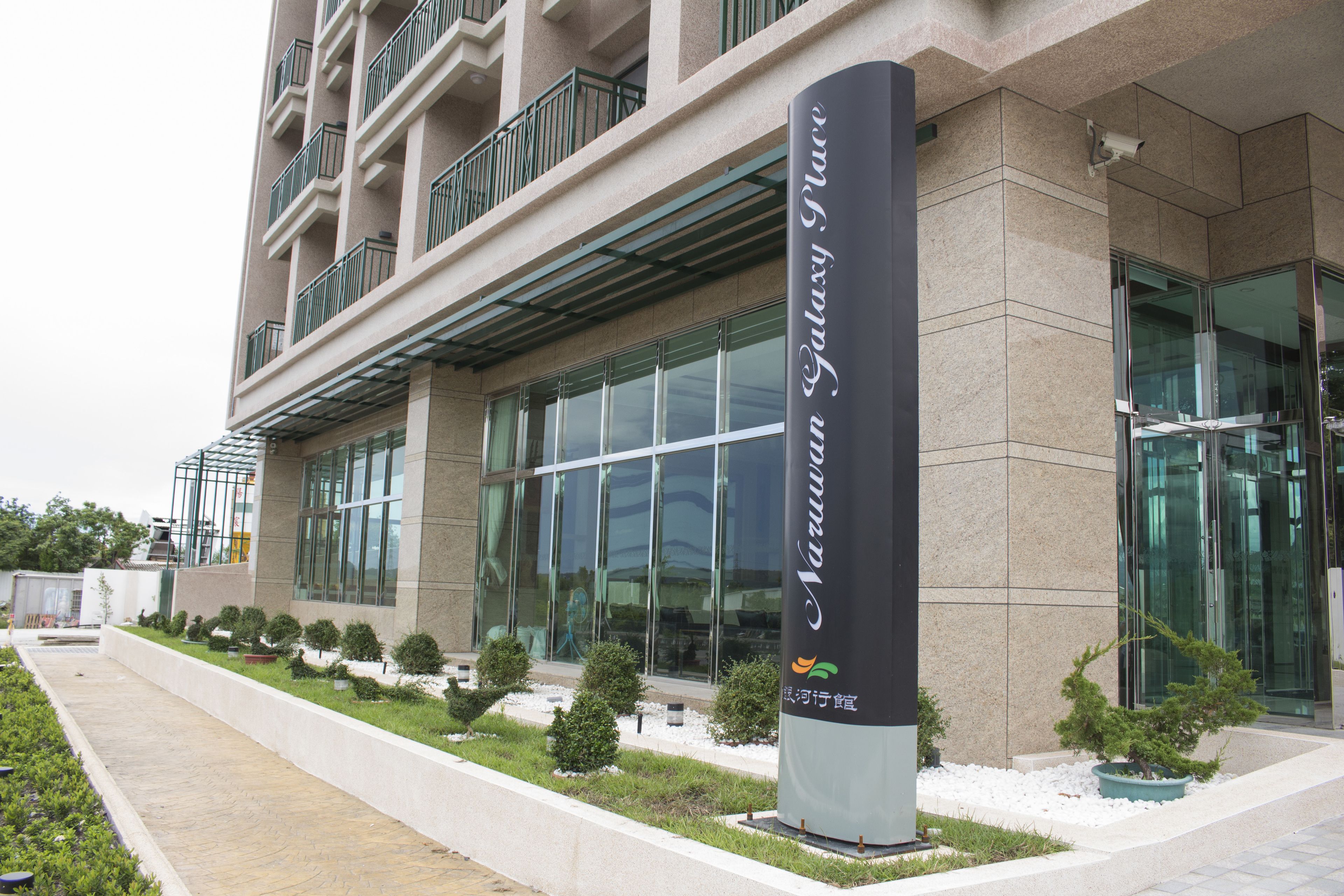 A modern building with a glass facade, a sign indicating 'Narrow Galaxy Place', and a landscaped area with small trees and shrubs.