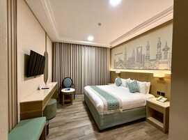 Habitación de hotel con cama, escritorio, silla, televisión, cuadro de un paisaje urbano y mesita de noche con teléfono.