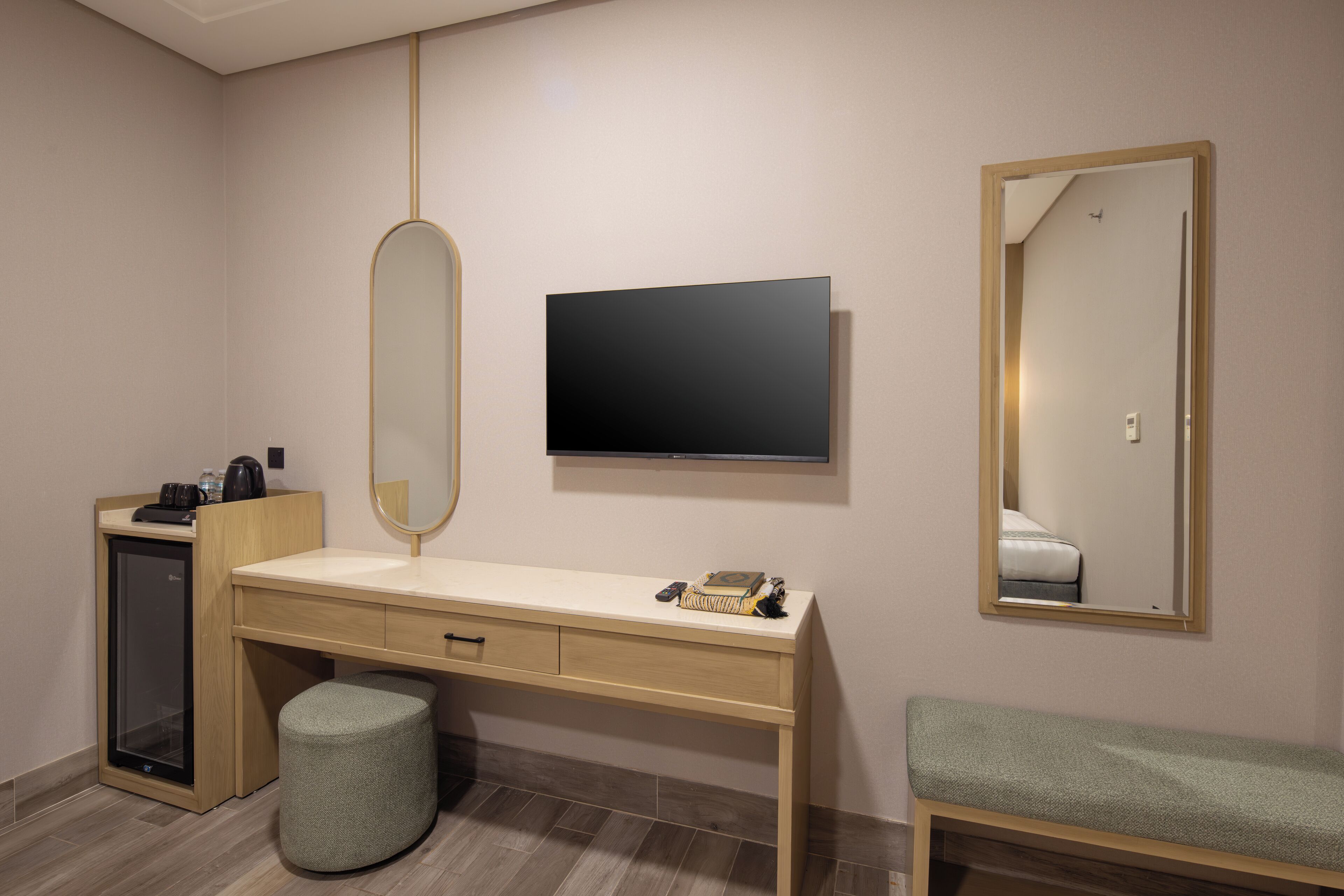 A hotel room with a wooden desk, a flat-screen TV, a mirror, a bench, and a minibar.