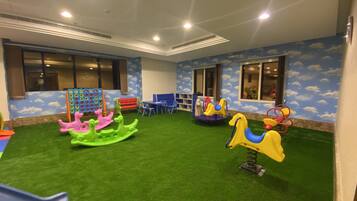 Zona de juegos infantiles con césped artificial, juegos de colores y un mural con cielo azul en las paredes.