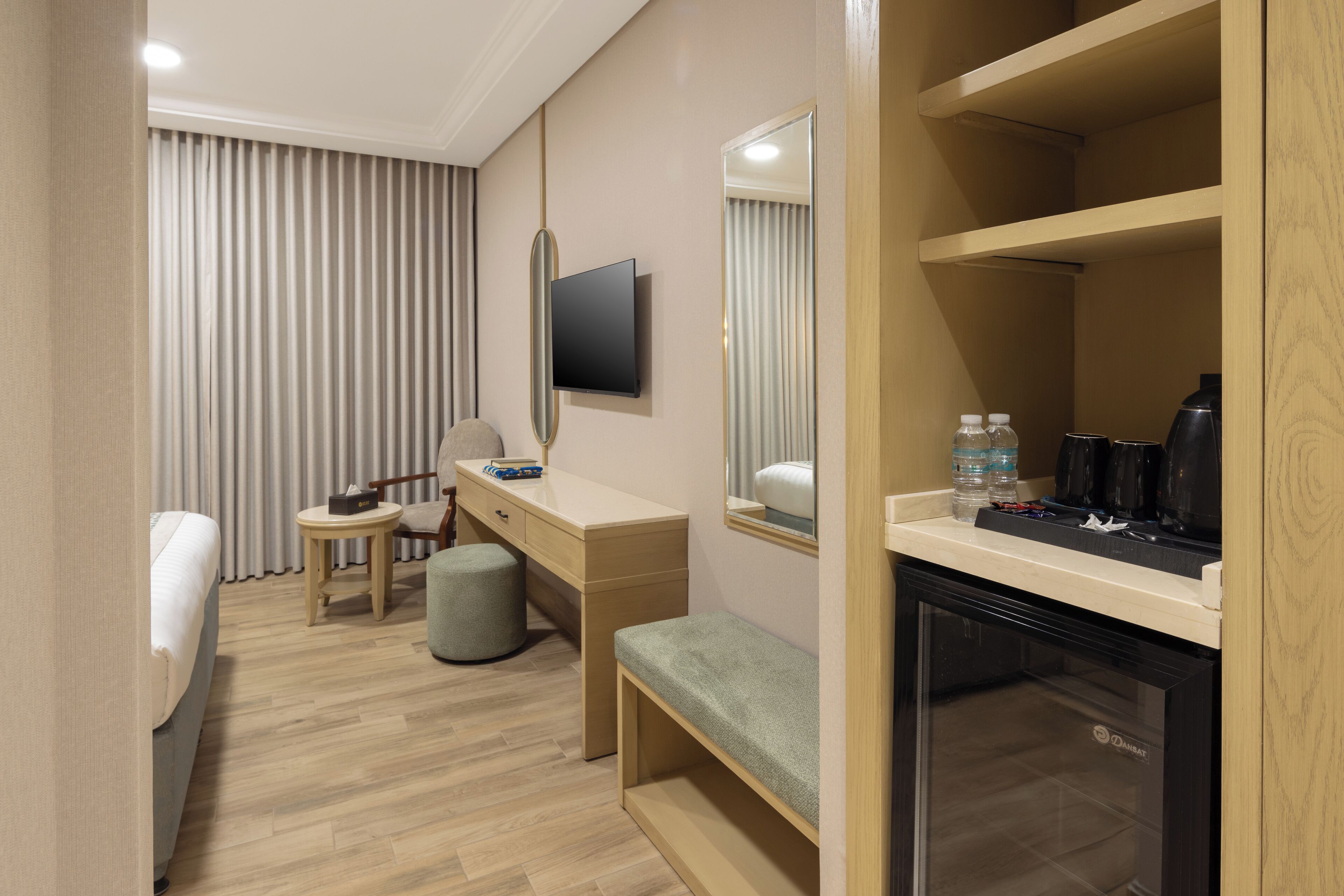 Een hotelkamer met een bed, een bureau, een stoel, een klein rond tafeltje, een bankje, een aan de muur bevestigde televisie, een spiegel en een minibar.