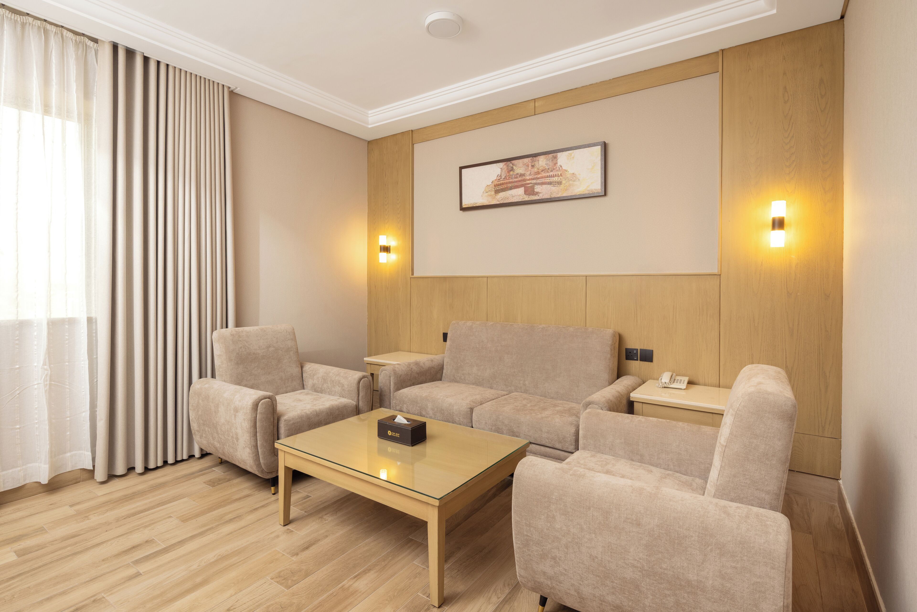 A modern hotel room with a beige sofa set, a glass coffee table, and a wooden floor. There are two wall-mounted light fixtures and a framed picture above the sofa.