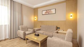 A modern hotel room with a beige sofa set, a glass coffee table, and a wooden floor. There are two wall-mounted light fixtures and a framed picture above the sofa.