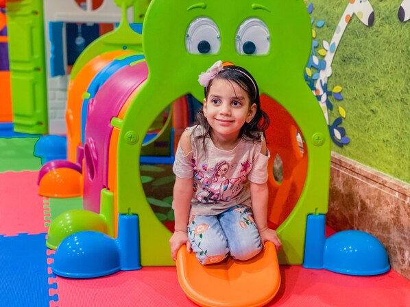 A child sitting on a colorful slide in a play area with a green slide structure and foam mats on the floor.
