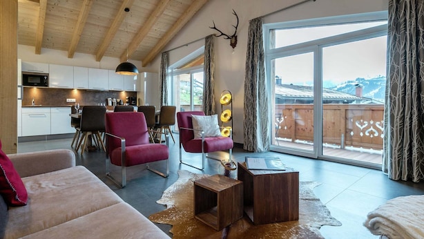 A spacious living area with a wooden ceiling, a large window with curtains, a dining area, and a kitchen.