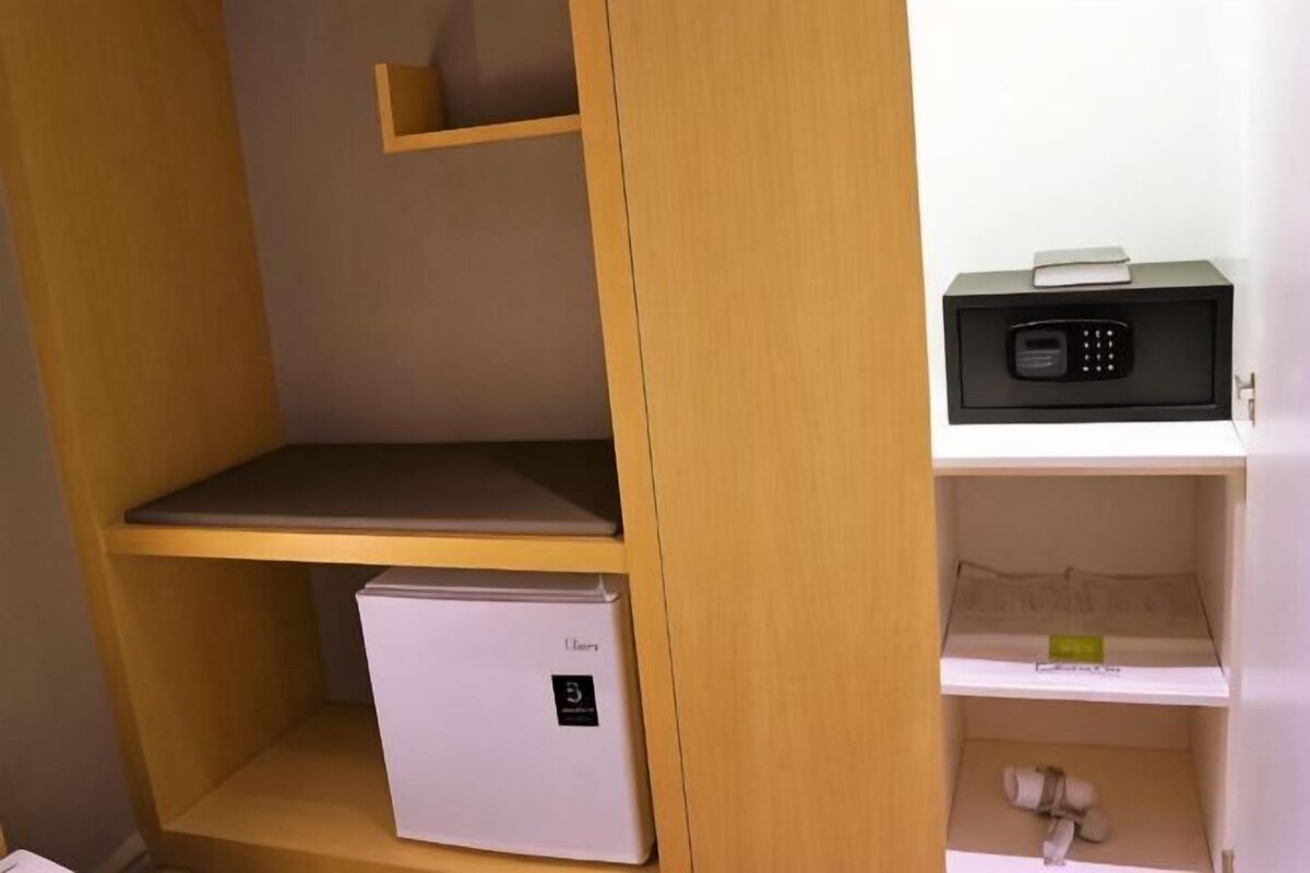 Habitación compacta con un mini refrigerador blanco, un dispositivo electrónico negro sobre una estantería y un mueble estante de madera.