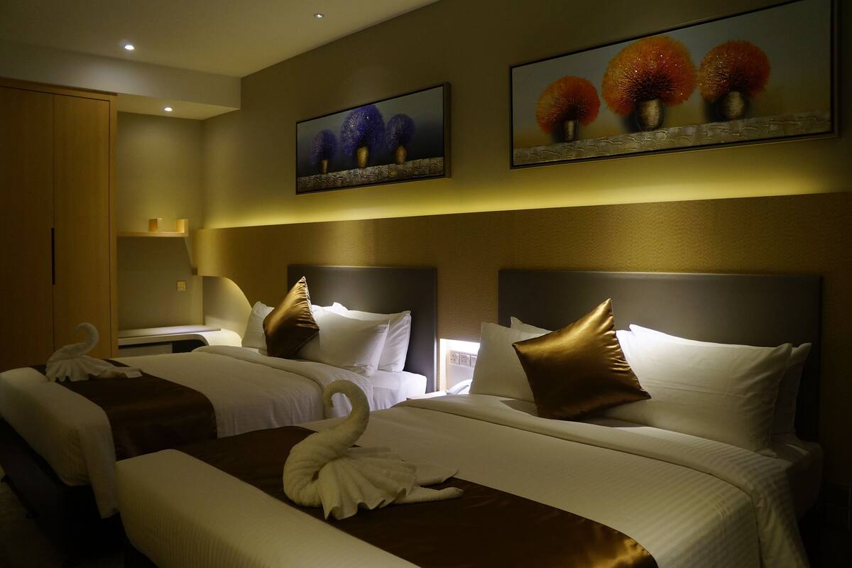 A hotel room with two beds, each with white linens and decorative pillows. There are wall-mounted paintings above the beds, and a wooden wardrobe to the left.