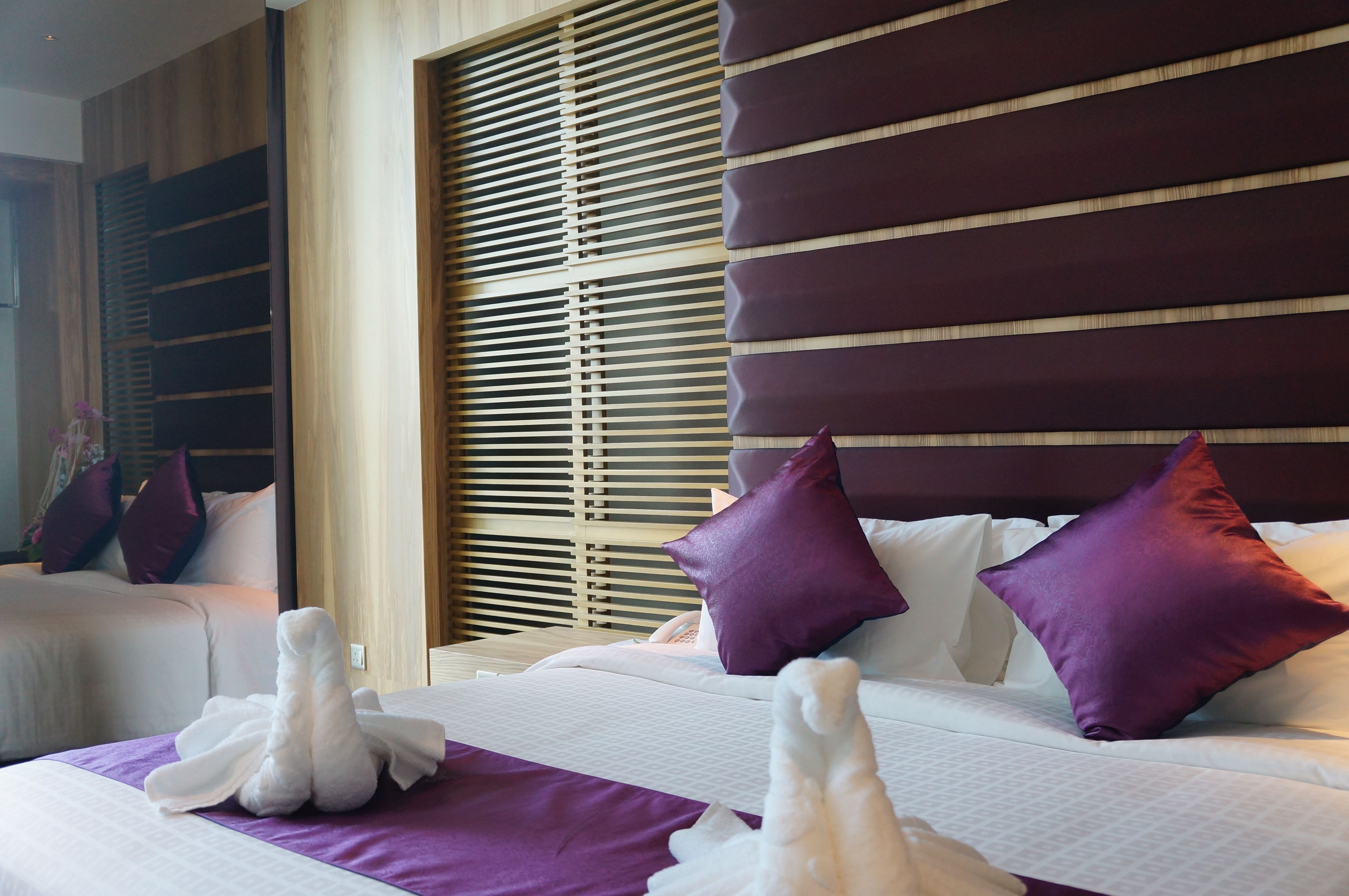 Une chambre d’hôtel avec un lit, des oreillers violets et une fenêtre avec des stores.
