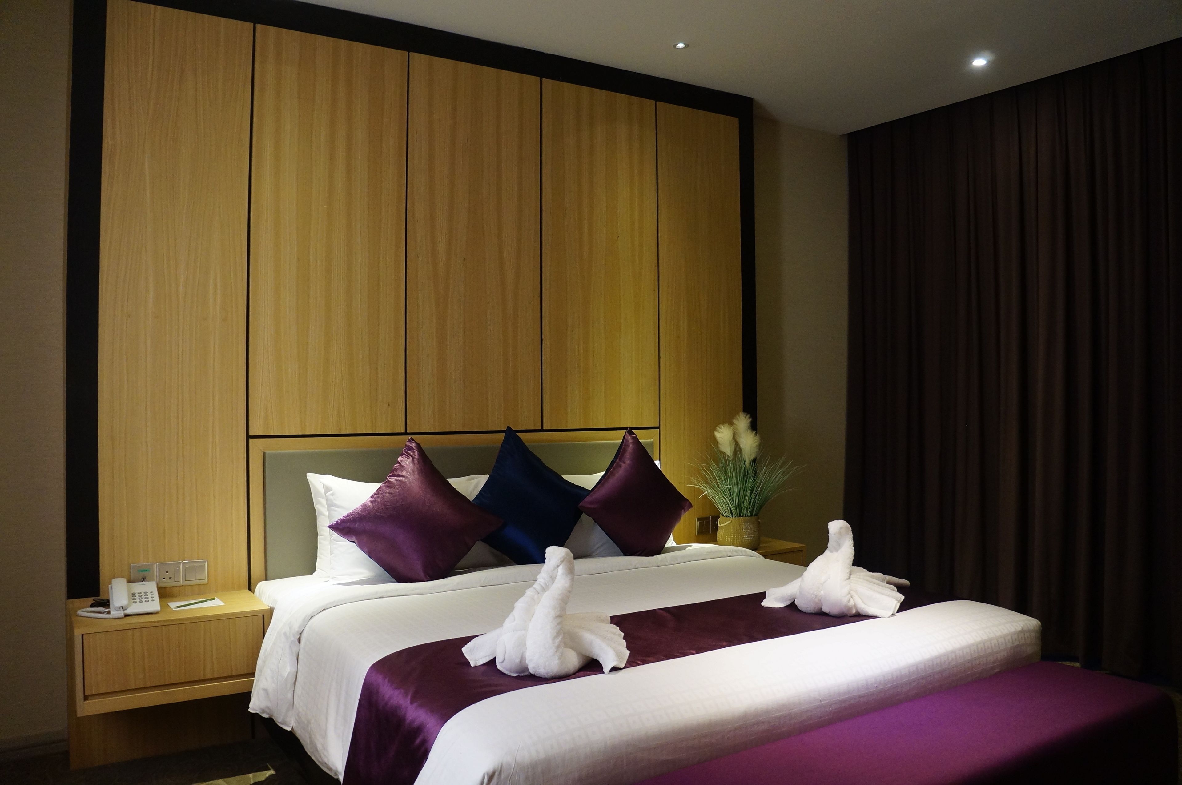 Een hotelkamer met een groot bed, twee kussens, een nachtkastje met een telefoon en een vaas met bloemen.