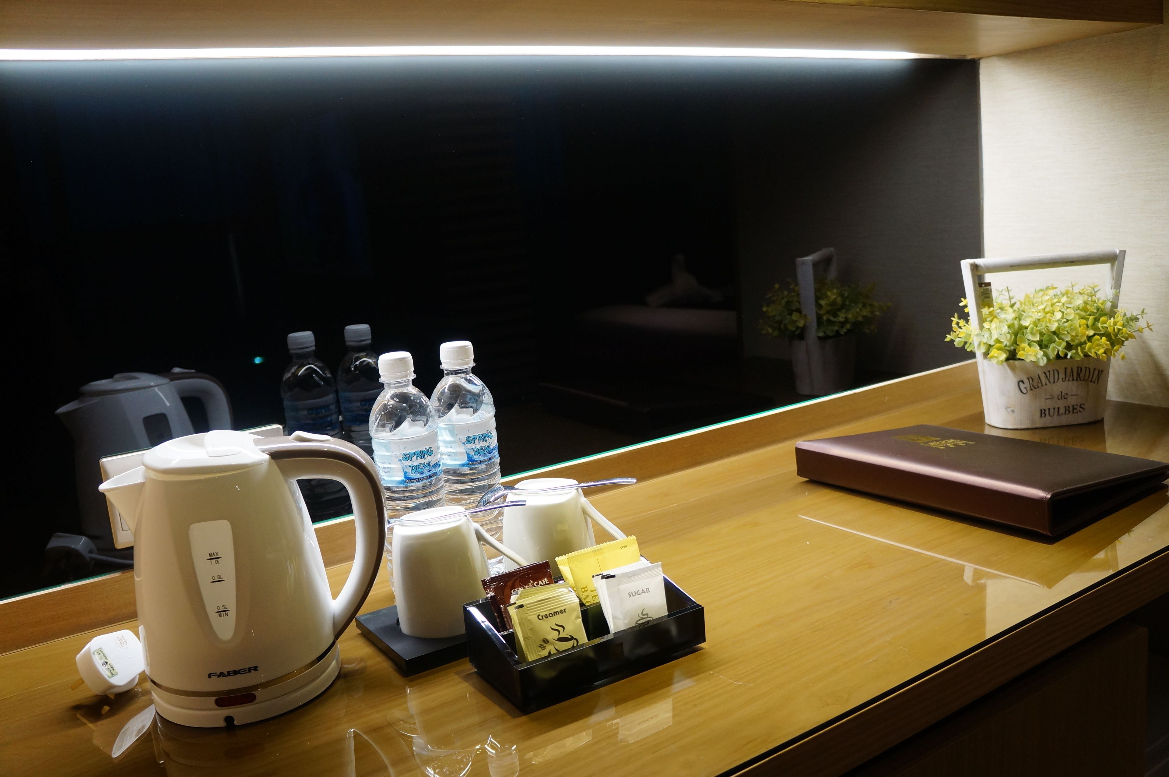 Zona de recepción de un hotel con cafetera, botellas de agua, bandeja con té y sobres de azúcar, y una planta decorativa.