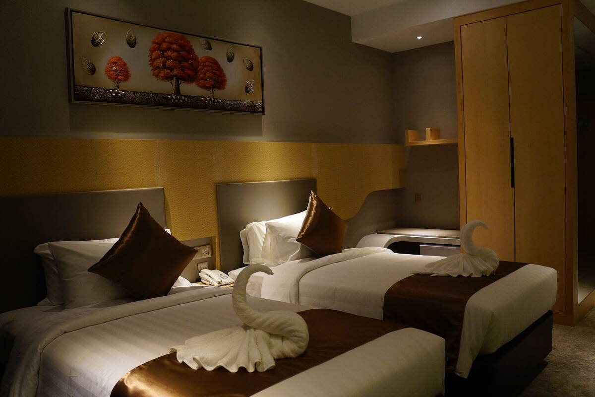 A hotel room with two beds, each with a brown headboard, white bedding, and a decorative swan towel. A painting of trees is on the wall above the beds.