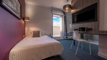 A modern hotel room with a bed, a desk with a flat-screen TV, a chair, and a large window offering a view of the sky.