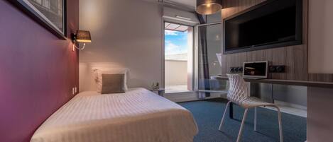 A modern hotel room with a bed, a desk with a flat-screen TV, a chair, and a large window offering a view of the sky.