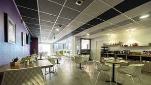 A modern dining area with multiple tables and chairs, a kitchen area with appliances and shelves, and a view of the city through large windows. - Nemea Appart Hotel Quai Victor Tours Centre (Tours)