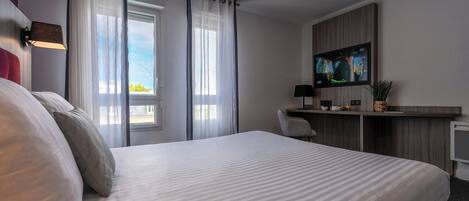 A hotel room with a bed, a desk with a TV, a window with curtains, and a plant.
