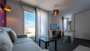A modern hotel room with a sofa, a small table, a desk, and a chair. There is a large window with curtains, a ceiling light, and a framed picture on the wall. - Nemea Appart Hotel Quai Victor Tours Centre (Tours)