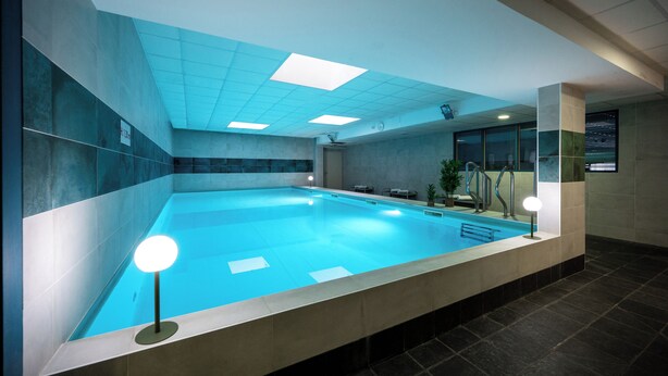 An indoor swimming pool with a tiled floor, a changing room with a bench, and a wall-mounted light fixture. - Nemea Appart Hotel Quai Victor Tours Centre (Tours)