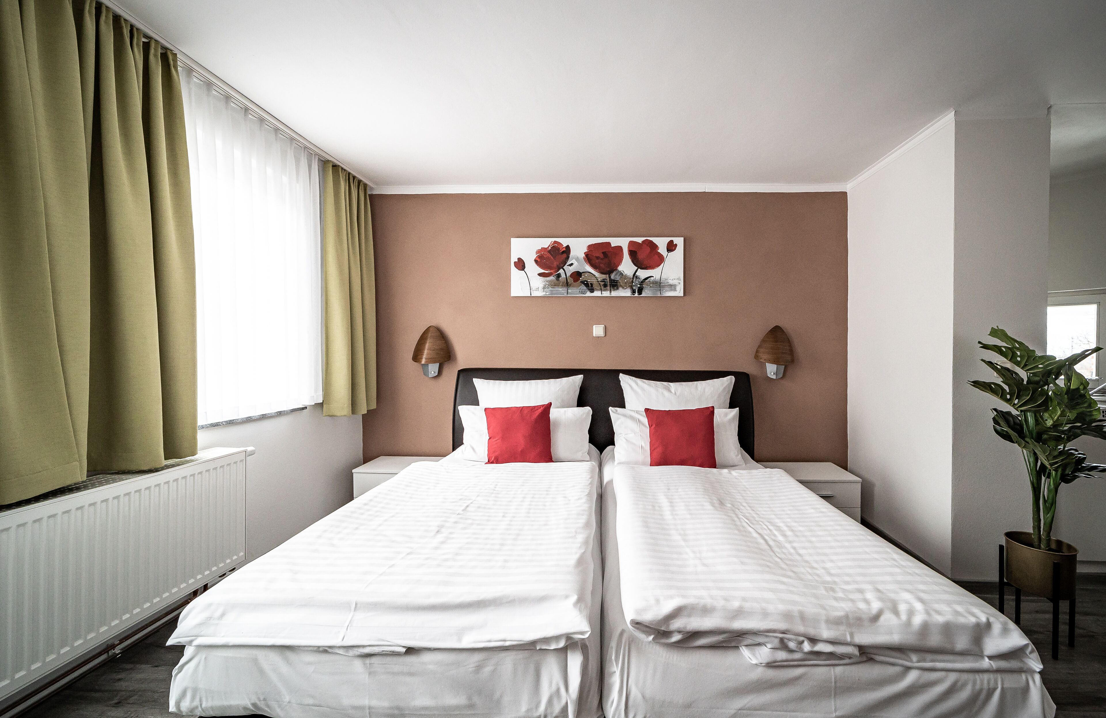 A hotel room with two single beds, a radiator, a window with curtains, a wall-mounted artwork, and a potted plant.